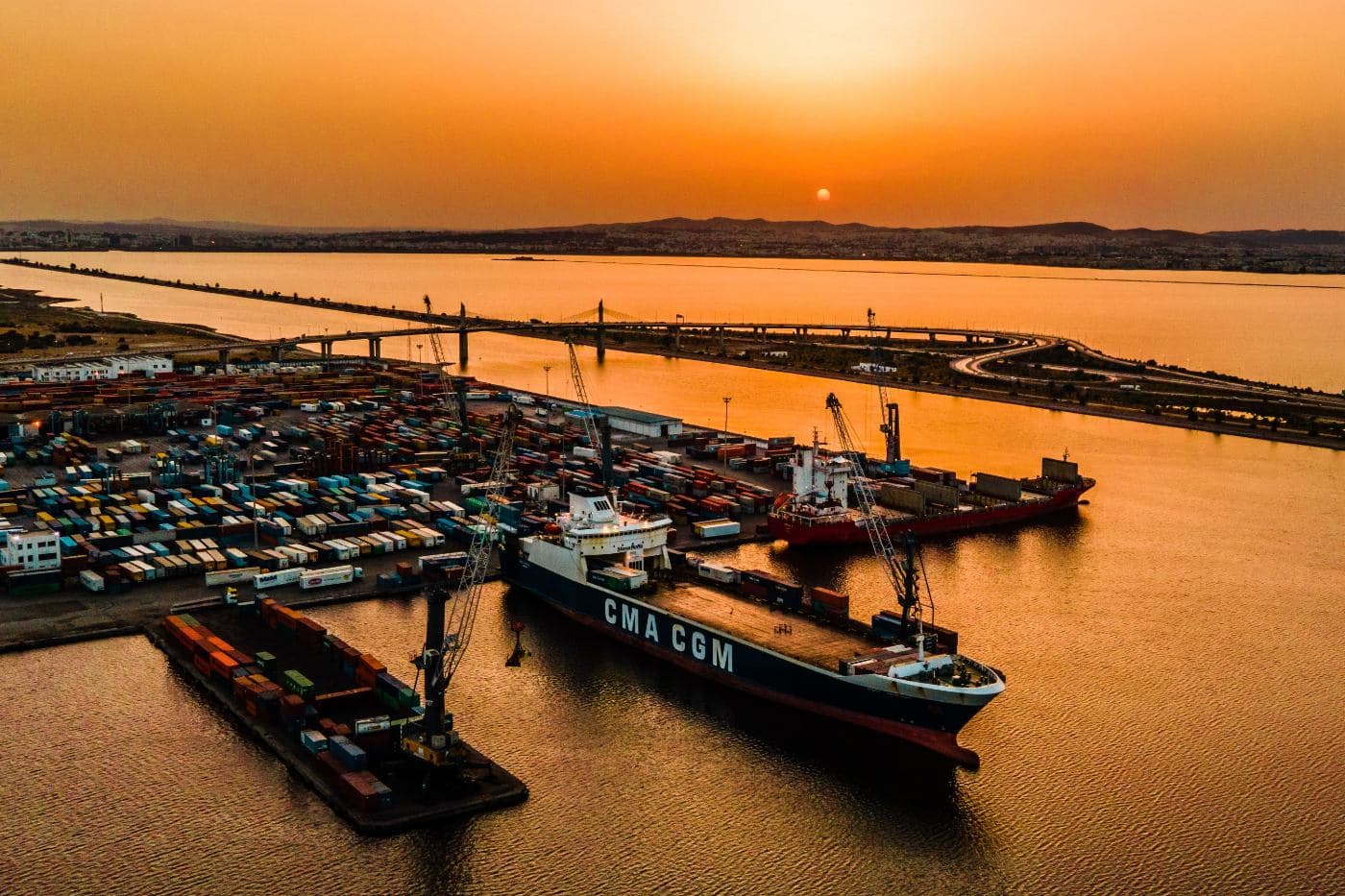 Un porte-conteneurs à quai au port de Radès, à Tunis, au crépuscule, avec le parc à conteneurs le long du quai et le pont routier qui enjambe le canal à l’arrière-plan.